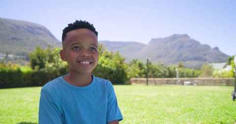 Smiling boy on sunlit grassy lawn with mountain view wearing blue t-shirt, joyful outdoor