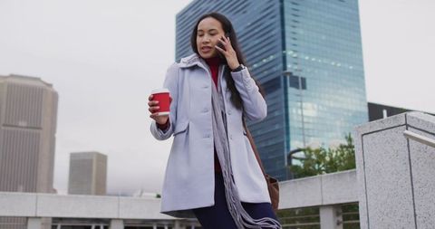 Businesswoman talking on phone while walking with coffee in downtown plaza
