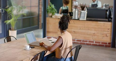 African american woman working remotely at communal cafe table with laptop and documents
