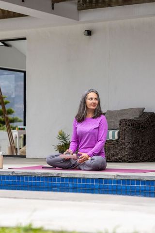 Mature woman sitting cross-legged on pink yoga mat at poolside terrace holding smartphone