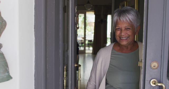 Senior woman with grey hair opening door at her home, dressed in casual attire, smiling warmly. Useful for themes related to hospitality, home visits, elderly trust, and family warmth.