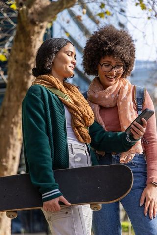 Young friends sharing phone while holding skateboard on college campus walkway