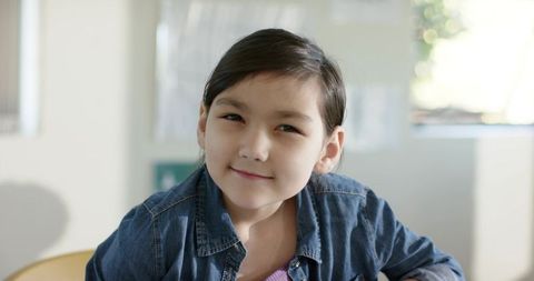Smiling asian girl sitting by window wearing denim jacket and pink top in bright room