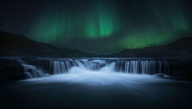 Midnight waterfall flowing beneath glowing green aurora borealis over rugged rock canyon
