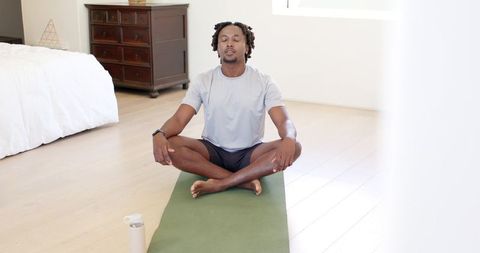 African american man meditating on green yoga mat in bright bedroom wearing wrist tracker