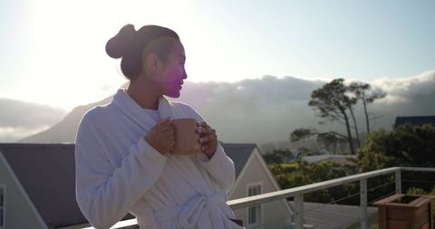 Asian woman standing on balcony in white bathrobe holding mug at sunrise mountain view
