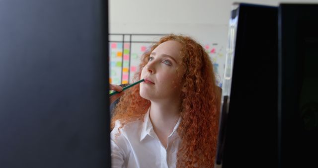 Young woman with red curly hair in professional office setting, appearing thoughtful while holding pen against her lips. Ideal for themes of brainstorming, strategic planning, professional concentration, and modern workspace. Useful for business, career, and motivational content on websites, presentations, and advertisements.