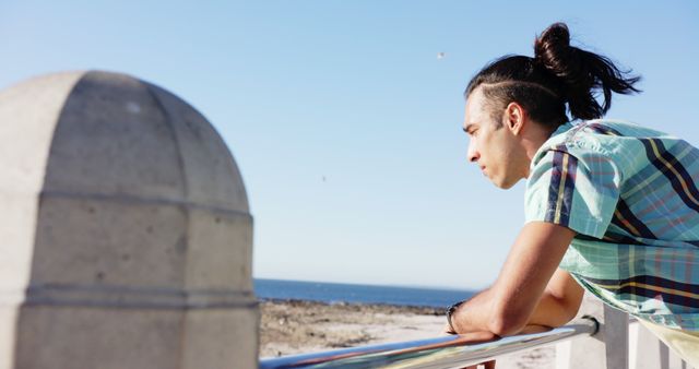 A young man with a man bun is looking out at the ocean, leaning on a rail, on a sunny day. The image captures a moment of contemplation, great for relaxation, tranquility, and self-thought concepts or outdoor activity-related content.