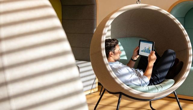 Executive reclining in round lounge pod analyzing tablet charts in modern coworking hub