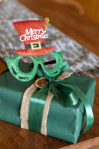 Wrapped green gift wearing glitter elf glasses and red merry christmas top hat with burlap ribbon