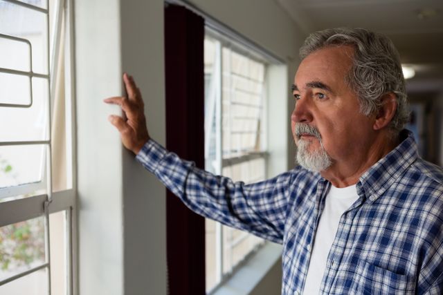 Thoughtful senior man looking through window while standing in nursing home