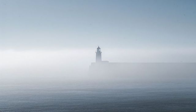 Solitary lighthouse emerging from dense fog over calm sea, misty coastal beacon at pier