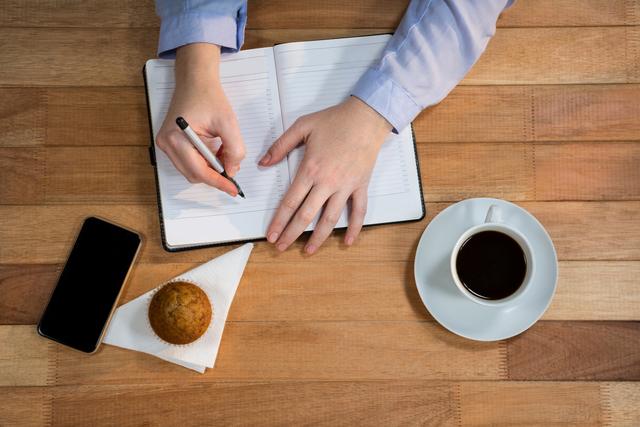 Businesswoman writing in a personal organizer at a wooden desk with a cup of coffee and a muffin. Ideal for use in articles or advertisements related to productivity, business planning, office routines, or professional organization. Can also be used in blogs about morning routines, effective work habits, or business tips.