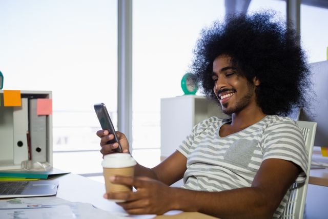 Smiling young man using phone while having coffee at desk in creative office