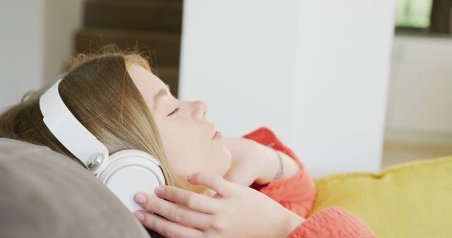 The teen girl is enjoying a quiet moment lounging on a couch, listening to music with her white headphones. The focus on her face shows her deeply immersed in the audio experience. Perfect for marketing home and lifestyle products, advertising audio and tech gadgets, or illustrating concepts of relaxation and enjoyment at home.