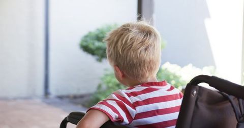 Young boy sitting in wheelchair on sunlit porch looking toward garden, calm reflective moment