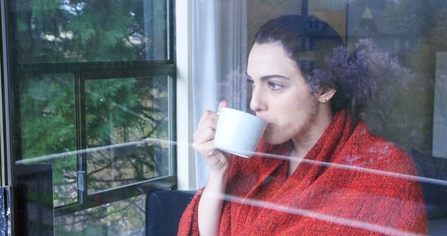 Woman covered with red blanket drinks coffee by window, looking outside. Morning light and greenery visible through glass. Perfect for themes of relaxation, contemplation, daily rituals, and cozy mornings.