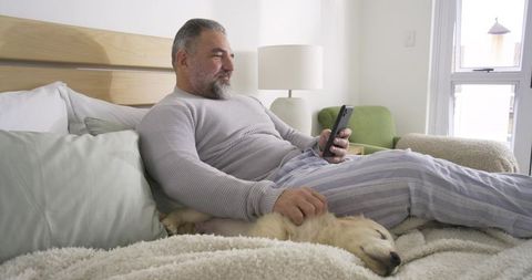 Man relaxing in bedroom with smartphone and golden retriever puppy