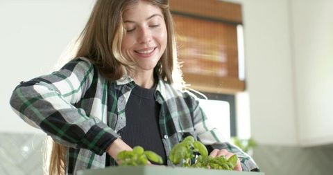 Smiling woman tending indoor herb planter on sunlit modern kitchen countertop