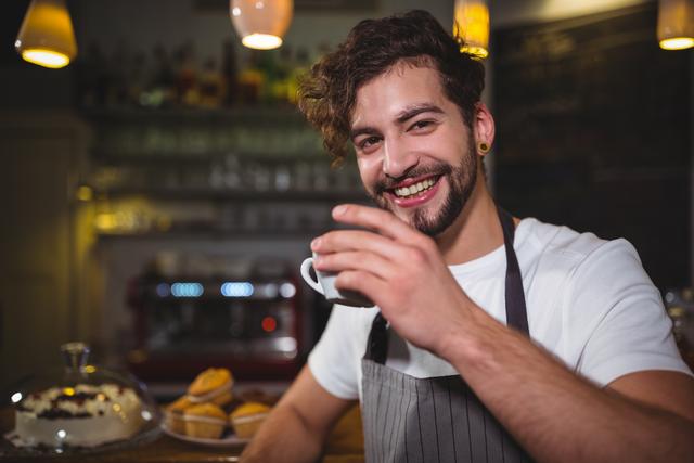 A young male waiter is smiling while enjoying a cup of coffee at the café counter. He is wearing an apron and appears relaxed and cheerful. This image can be used for promoting café culture, hospitality services, or small businesses. It is ideal for websites, advertisements, and social media posts related to coffee shops, baristas, and customer service.