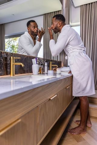 African american man applying facial cream at luxury bathroom vanity wearing white robe