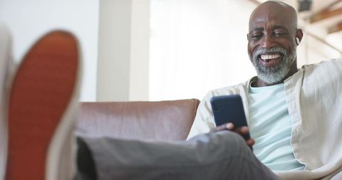 Senior african man relaxing on sofa, smiling while using smartphone and earbud