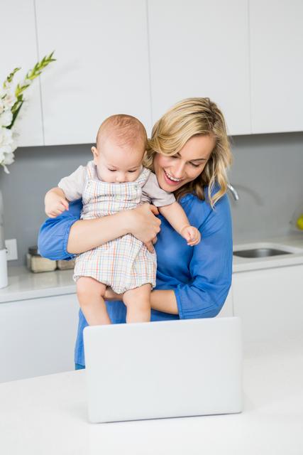 Mother holding baby boy while using laptop in modern kitchen. Ideal for themes related to parenting, multitasking, work-from-home, family life, and modern technology. Can be used in articles, blogs, advertisements, and social media posts about work-life balance, motherhood, and home life.