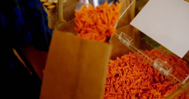 This image depicts the process of filling a brown paper bag with uncooked red pasta from a bulk bin. It illustrates themes of eco-friendly shopping, sustainability, and zero waste principles. Perfect for use in articles or content related to eco-conscious lifestyle choices, bulk food shopping, and plastic-free grocery supplies.