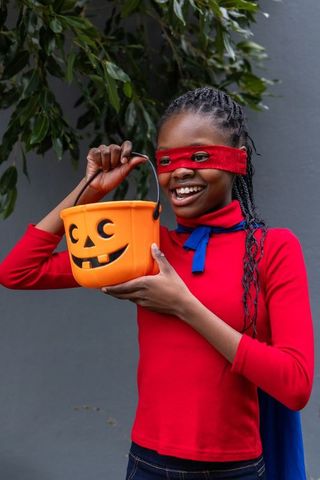 Girl in red mask and blue cape holding jack-o-lantern bucket while smiling outdoors