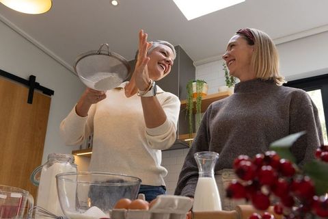 Senior woman and friend baking together, sifting flour in cozy modern kitchen