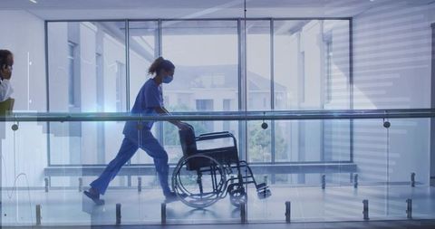 Nurse pushing wheelchair down bright hospital corridor with glass railings
