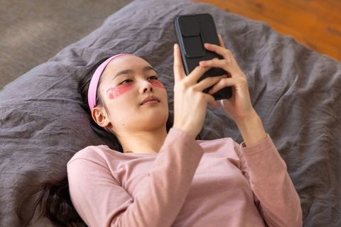 Relaxed woman practicing skincare and browsing phone in bedroom