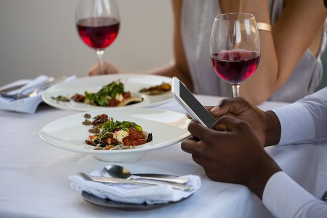 Couple enjoying a meal at a restaurant with wine glasses and gourmet dishes on the table. Man using a smartphone, indicating modern communication and social media usage. Ideal for illustrating themes of dining out, romantic dinners, technology in daily life, and social interactions.