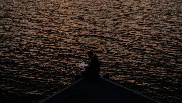 Sitting solitary man reading on pointed dock at dusk silhouetted by rippling amber water