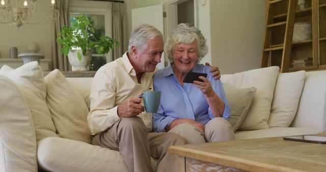 Senior couple sitting on couch, smiling while looking at smartphone. Man holding a mug. Cozy living room setting with plants and wooden furniture. Suitable for concepts like technology use among the elderly, relaxing at home, happy retirement, and bonding moments.