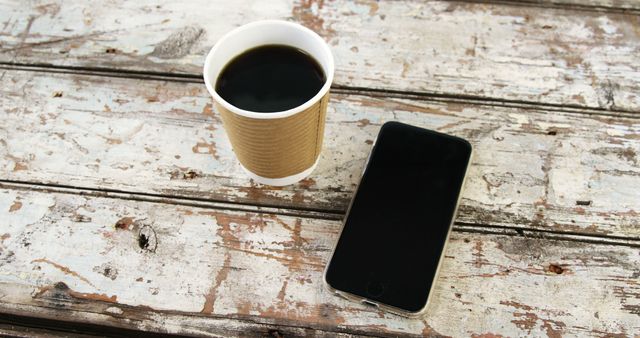 A disposable coffee cup next to a smartphone on a rustic wooden table, with copy space. These items suggest a break or work setting, where someone might be enjoying a beverage while staying connected.