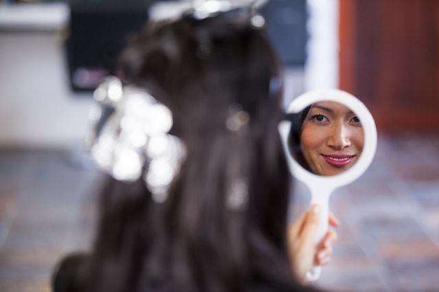 Woman with hair dye looking at her reflection in a hand mirror at a salon. Ideal for use in beauty and hair care advertisements, salon promotions, self-care articles, and personal grooming blogs.