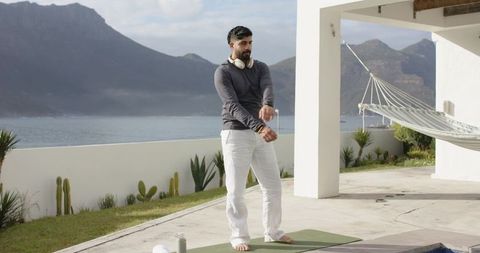 Indian man adjusting sleeves by pool on yoga mat wearing headphones overlooking sea and mountains