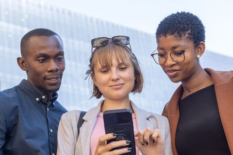 Diverse young professionals collaborating over smartphone outside modern glass office building