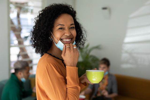 African American woman smiling while holding a cup of coffee and pulling down her facemask. Colleagues in the background sitting on a couch, talking during a coffee break. Ideal for use in articles about workplace culture, office breaks, team bonding, and modern office environments.