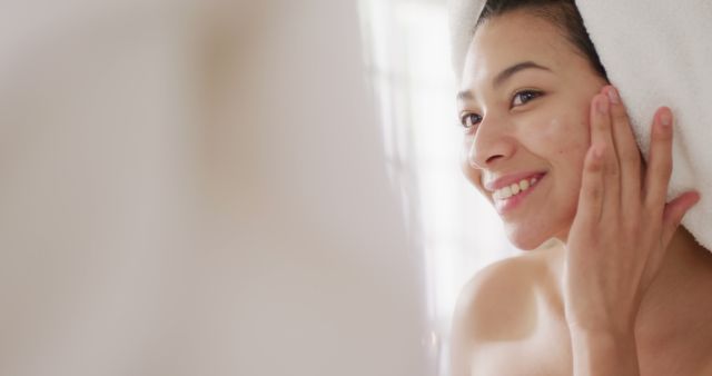 Woman smiling while applying skin care regimen in bathroom, wrapped in towel. Ideal for promoting beauty products, self-care routines, and healthy skin tips. Useful for blogs, advertisements, and wellness content.