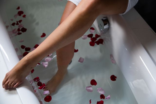Close up the legs of a young Caucasian woman, with one foot in the water with rose petals on the side, while she sits on the side of the bathtub