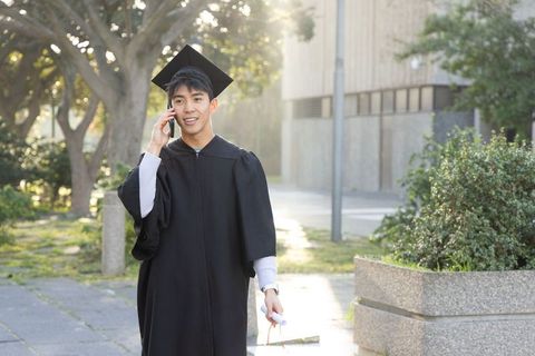 Asian graduate walking campus path talking on smartphone holding diploma in warm sunlight