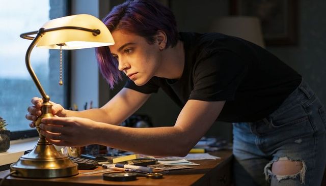 Young woman adjusting brass desk lamp at cluttered creative workspace by window, concentrating