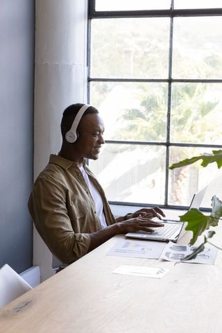 Mid adult African American man typing on laptop by window wearing headphones, remote work
