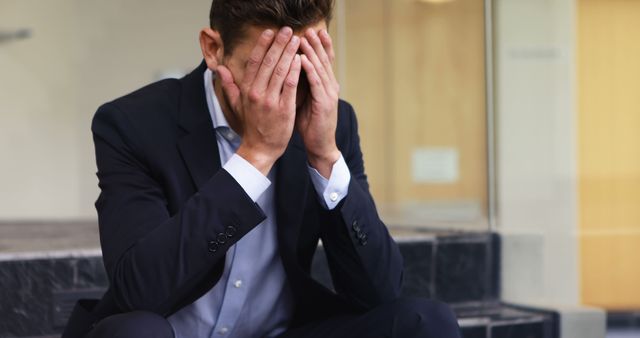 A young Caucasian businessman appears stressed or upset, covering his face with his hands, with copy space. His posture and expression convey a sense of disappointment or emotional distress, related to his professional life.