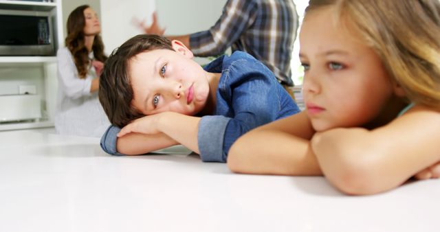 Image shows two children looking sad while sitting at a table, with out-of-focus parents arguing in the background. Great for articles or content on family conflicts, emotional effects on children, or domestic issues. Can be used in online publications, psychological studies, or family counseling resources.