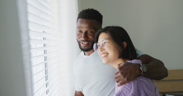 Happy diverse couple embracing and looking through window in bedroom - Download Free Stock Photos Pikwizard.com