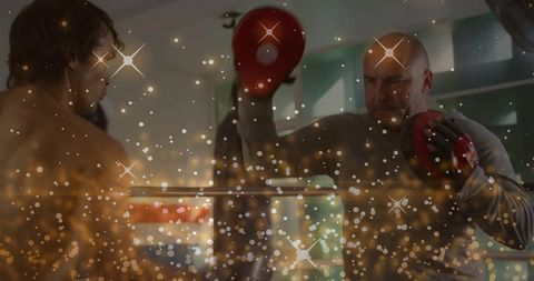 Boxer punching while coach holds red focus mitts in gym with sparkling bokeh lights