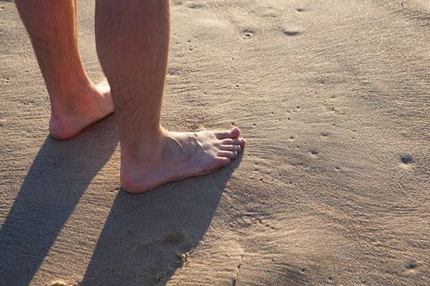 Standing barefoot on sunlit beach sand, close-up of lower legs and toes casting shadow